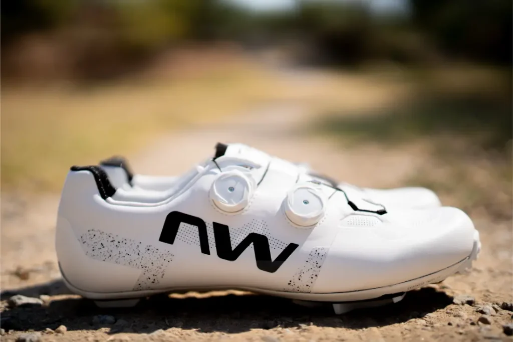 Un par de zapatillas de ciclismo Northwave Extreme X blancas, con el logotipo «NW» en negro y dos cierres de dial, colocadas en un camino de tierra al aire libre, que ofrecen un rendimiento de gama alta y una versatilidad excepcional para los ciclistas más exigentes.