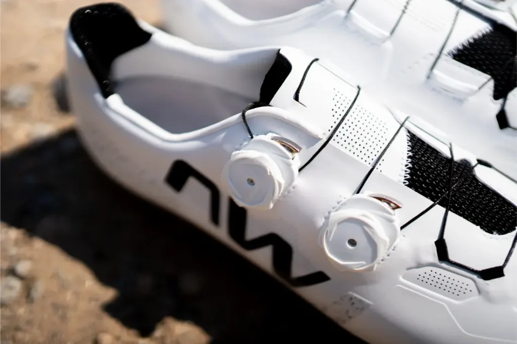 Primer plano de un par de zapatillas de ciclismo blancas con detalles en negro y cierres de dial, colocadas sobre una superficie rugosa al aire libre. Las Northwave Extreme X destacan en esta imagen, lo que refleja su diseño de gama alta y su reconocida versatilidad para los ciclistas que buscan tanto rendimiento como estilo.