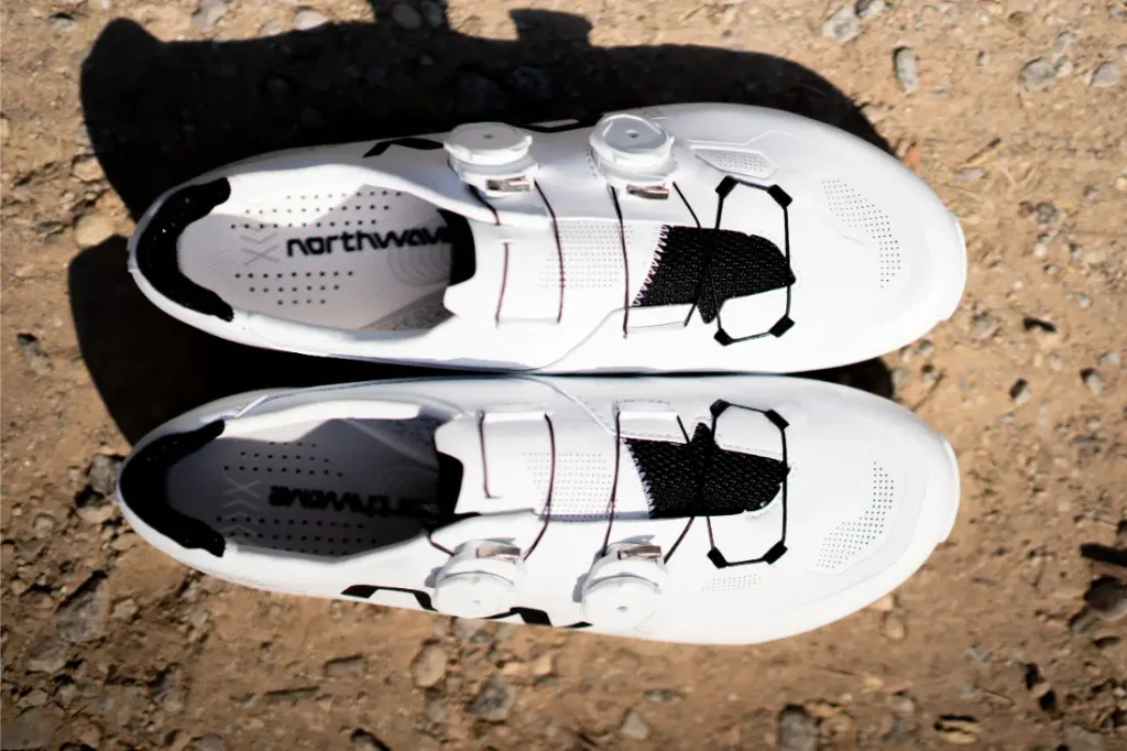 Un par de zapatillas de ciclismo Northwave Extreme X blancas, con malla negra y detalles geométricos, famosas por su versatilidad de alta gama, colocadas sobre una superficie de grava.