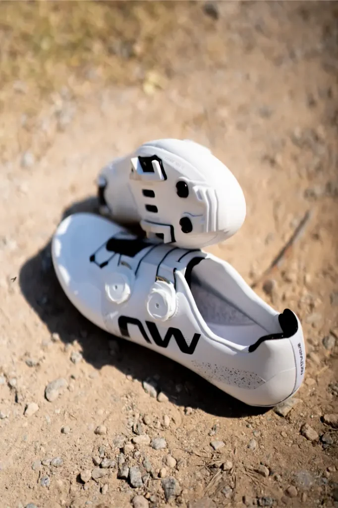 Un par de zapatillas de ciclismo Northwave Extreme X blancas, con detalles en negro y calas, están colocadas sobre un camino de tierra, con la luz del sol proyectando sombras, lo que pone de manifiesto su versatilidad para los ciclistas que buscan un rendimiento de gama alta.