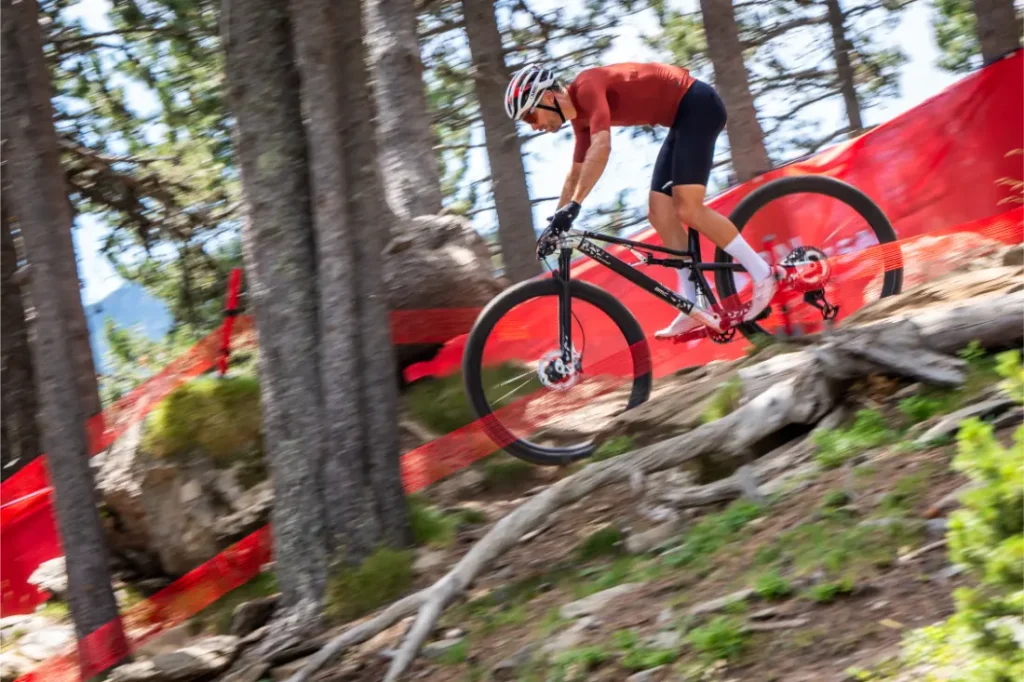 Un ciclista con casco baja en bicicleta de montaña por un sendero boscoso y rocoso, mostrando la «revolución ruedas» con ruedas de 32, mientras que al fondo se ve una red de seguridad roja.