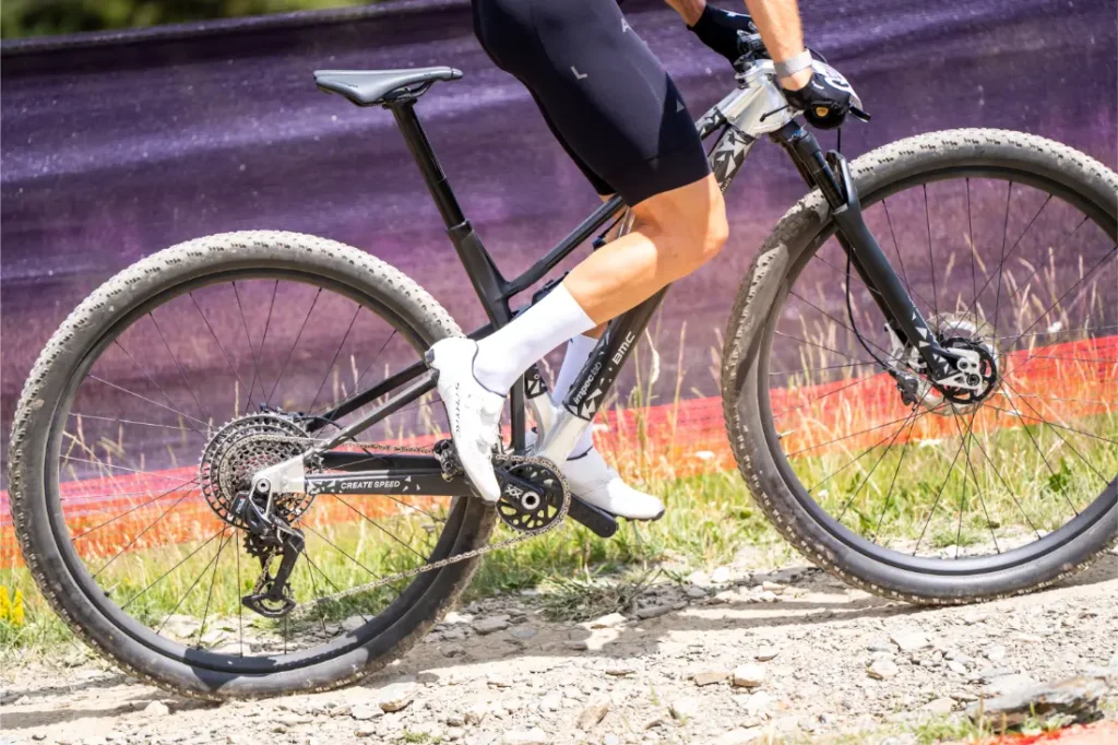 Un ciclista que lleva pantalones cortos negros y zapatillas blancas monta en una bicicleta de montaña con ruedas de 32 por un sendero de tierra rocoso, con una barrera morada y naranja al fondo.