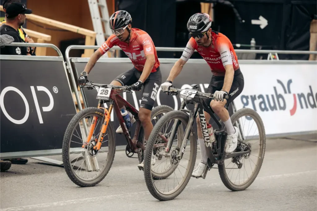 Dos ciclistas con maillots rojos y dorsales con los números 28 y 27 se detienen con sus bicicletas de montaña, equipadas con ruedas de 32, junto a una barrera metálica en un evento ciclista al aire libre, encarnando el espíritu de la revolución necesaria dentro de la industria.