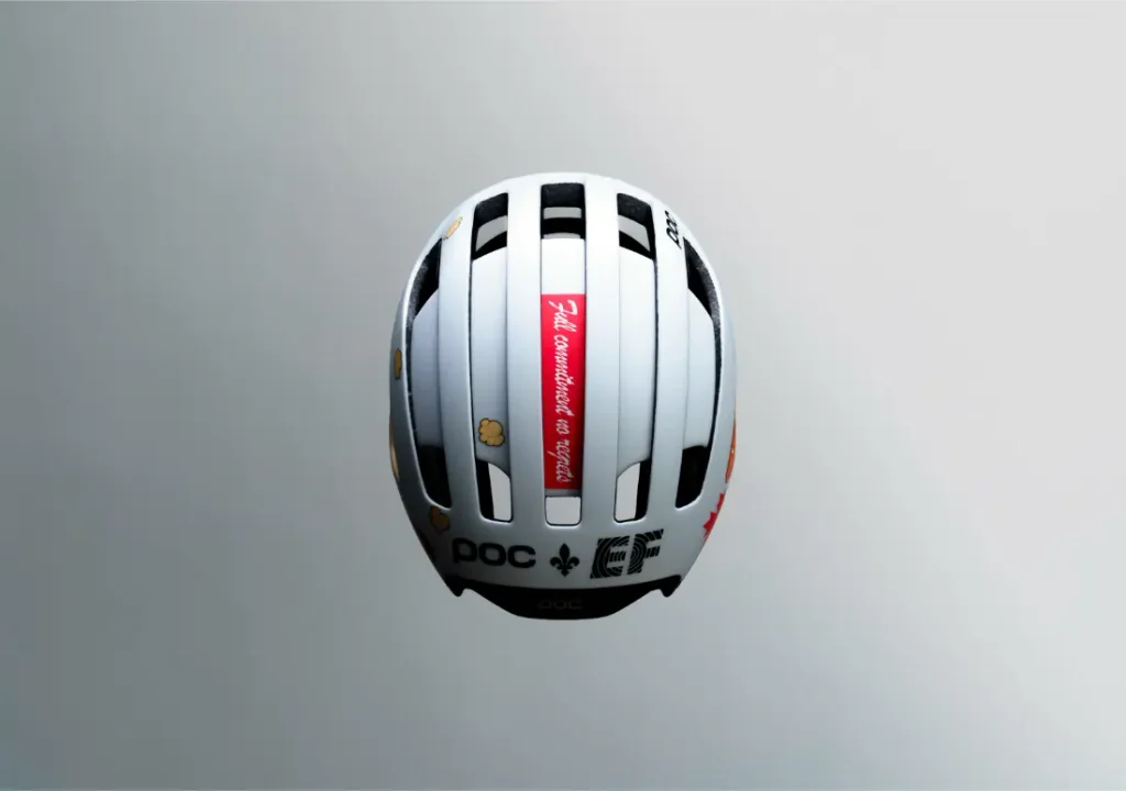 Casco de bicicleta blanco visto desde arriba, con rejillas de ventilación negras, varios logotipos y una etiqueta roja con texto en el centro.