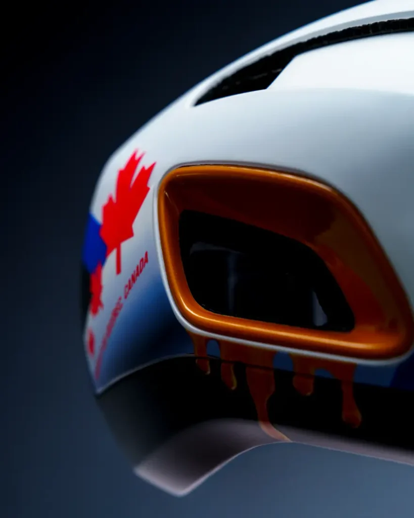Primer plano de un casco de carreras blanco con una rejilla de ventilación naranja, una hoja de arce roja y la inscripción «CANADA». También se aprecian detalles en azul y rojo en el casco.