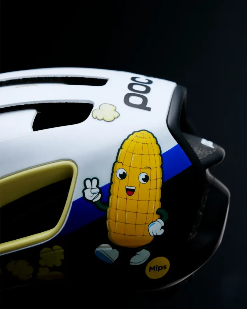 Un casco de bicicleta blanco y negro muestra una mazorca de maíz de dibujos animados con brazos, piernas y una cara sonriente, haciendo el signo de la paz, junto con pequeños dibujos de palomitas.