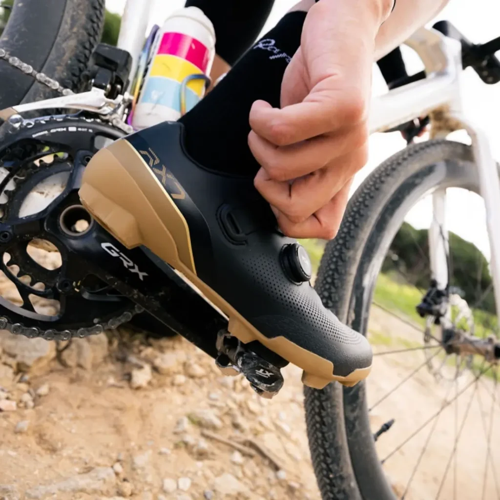 Una persona engancha unas zapatillas de ciclismo Shimano RX710, de color negro y marrón claro, a un pedal GRX en un camino de grava, haciendo gala de tecnología de competición, con un bidón visible en el cuadro de la bicicleta.