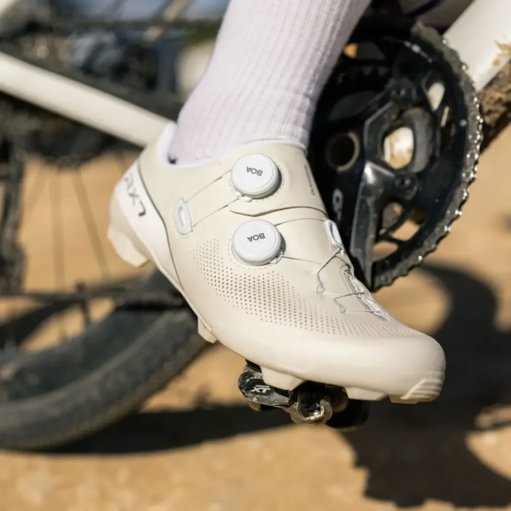 Primer plano de una persona que lleva unas zapatillas blancas de ciclismo Shimano RX710 enganchadas al pedal de una bicicleta, con parte del juego de bielas de la bicicleta y un camino de tierra visibles al fondo, lo que pone de relieve la tecnología Gravel para un rendimiento óptimo en terrenos accidentados.