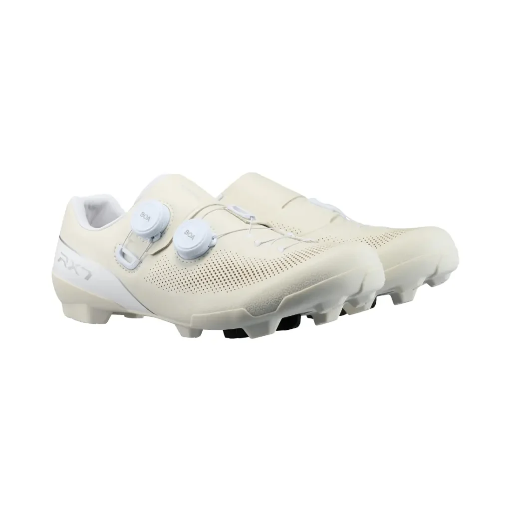 Un par de zapatillas de ciclismo Shimano de color blanco roto con parte superior perforada, dos cierres de dial blancos y suelas de goma texturizadas diseñadas para ofrecer agarre en senderos de grava, que incorporan tecnología de competición para un rendimiento óptimo.