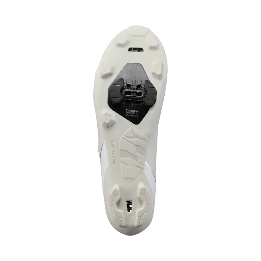 La parte inferior de unas zapatillas de ciclismo blancas Shimano RX710, en las que se aprecia una cala Shimano SPD fijada a la suela, con el dibujo de la banda de rodadura visible y las inscripciones «Carbon» y «Shimano», además de características inspiradas en la tecnología de competición para ofrecer un rendimiento superior en grava.