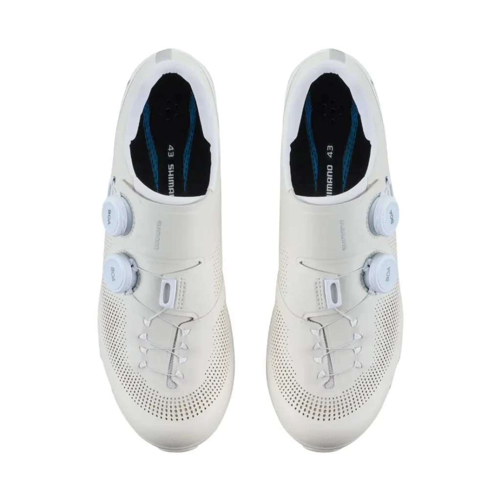 Un par de zapatillas de ciclismo blancas de Shimano con cierre de doble dial, parte superior perforada y plantillas negras, vistas desde arriba: perfectas para aventuras de gravel y repletas de tecnología de competición.