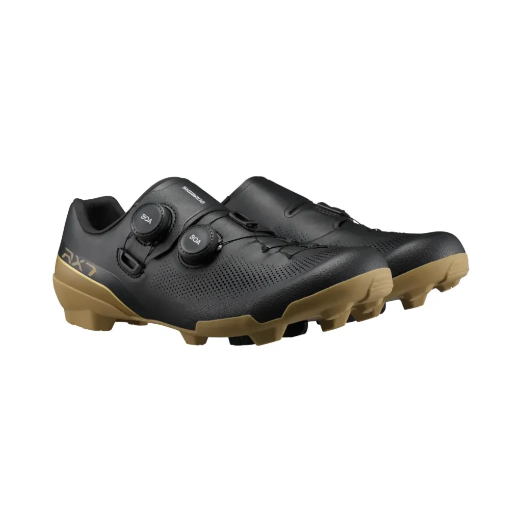 Un par de zapatillas de ciclismo Shimano RX710 negras con suela de goma de color beige, que cuentan con dos cierres de dial en cada zapatilla para un ajuste personalizable; diseñadas especialmente para salidas por grava y mejoradas con tecnología de competición para un rendimiento superior.