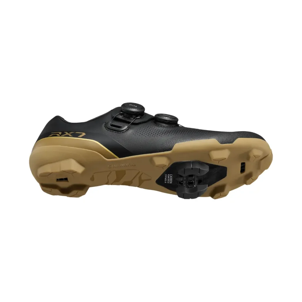 Se muestra una zapatilla de ciclismo negra con suela de color beige vista desde el lateral y desde abajo, que cuenta con una cala, una suela con tacos y un sistema de cierre de doble dial. Diseñadas para ciclistas de gravel, estas zapatillas Shimano incorporan tecnología de competición para ofrecer un rendimiento excepcional en terrenos variados.