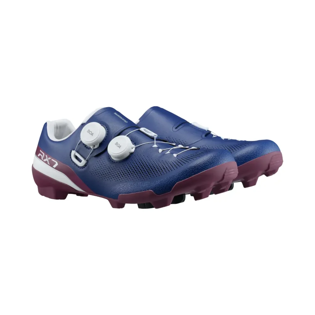 Un par de zapatillas de ciclismo Shimano RX710 azules con diales blancos y suelas granates, diseñadas con tecnología de competición para el ciclismo de grava, mostradas sobre un fondo blanco.