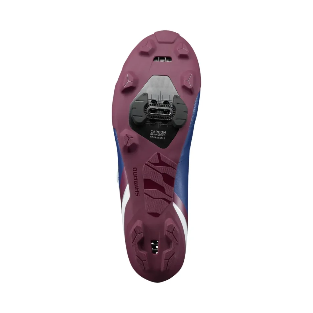 La suela de una zapatilla de ciclismo Shimano RX710, con un dibujo morado, una cala Shimano SPD y una placa de compuesto de carbono, vista desde abajo: diseñada a la perfección para salidas por grava e incorporando tecnología de competición.