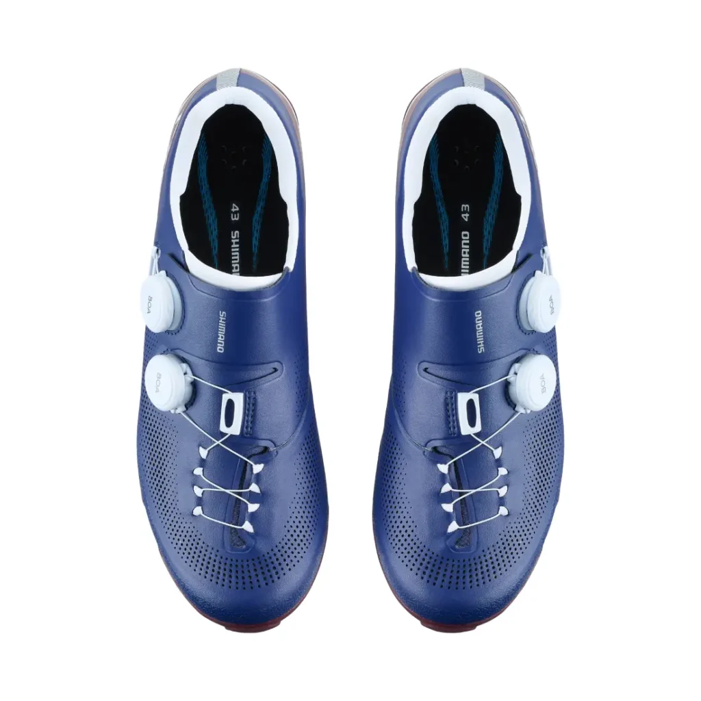 Un par de zapatillas de ciclismo Shimano RX710 azules con cierres de dial blancos y plantillas negras, diseñadas con tecnología de competición para el ciclismo de grava, vistas desde arriba sobre un fondo blanco.