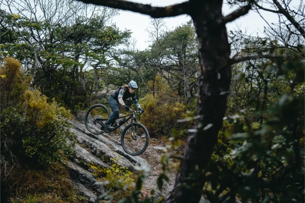 Una persona que lleva casco va en bicicleta de montaña con ruedas de 32 por un sendero rocoso y boscoso, rodeado de árboles y arbustos.