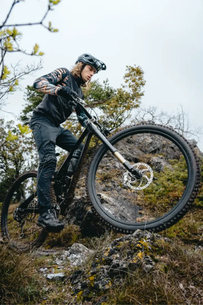 Una persona que lleva casco y ropa de montaña monta en bicicleta de montaña con ruedas de 32, subiendo por un terreno rocoso, rodeada de vegetación y árboles.