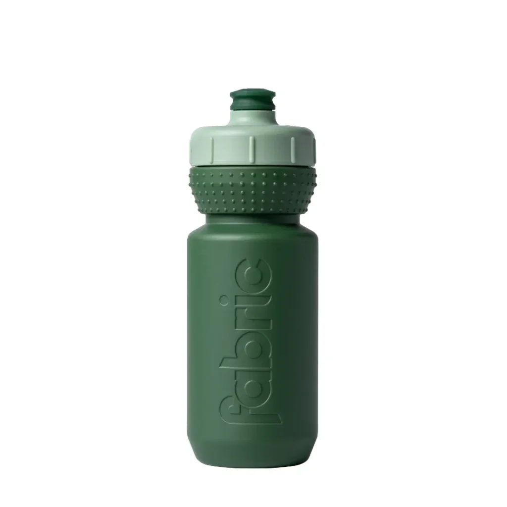 Botella de agua de plástico verde con tapón de rosca texturizado y la palabra «fabric» grabada en relieve verticalmente en el lateral, que cuenta con «bidón protección antimicrobiana» para una mayor higiene. El «Fabric Gripper V2» aparece representado sobre un fondo blanco.