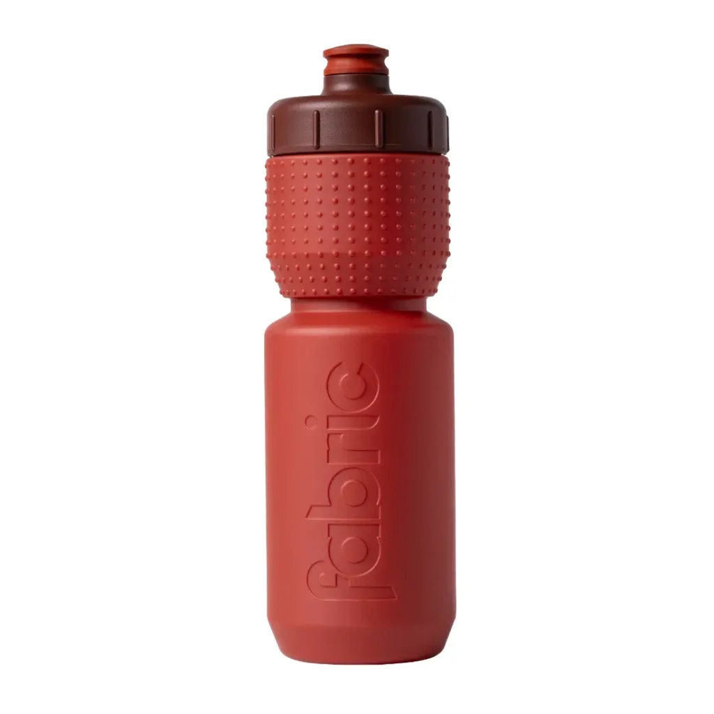 Botella de agua de plástico roja con un tapón de rosca marrón texturizado y la palabra «fabric» grabada en relieve verticalmente en el lateral, representada sobre un fondo blanco. Este bidón antimicrobiano Fabric Gripper V2 ofrece protección permanente para tus necesidades de hidratación.