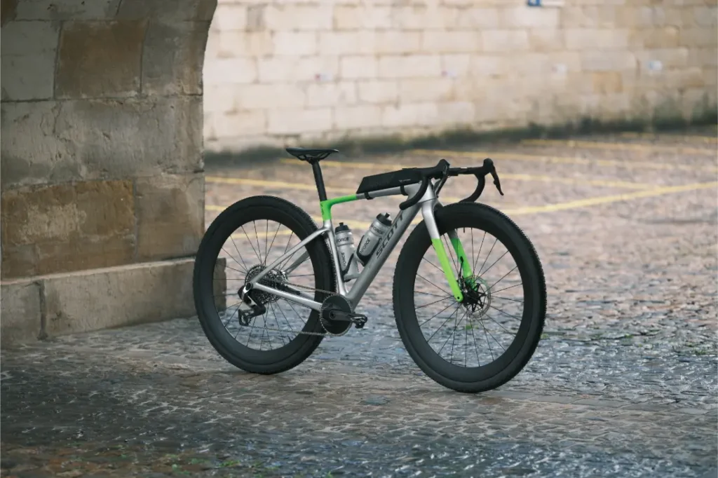 Una bicicleta de ruedas gruesas, de color plateado y verde, con manillar de carretera —símbolo de la «revolución de las ruedas» en la industria del ciclismo— está aparcada sobre el empedrado mojado, junto a un arco de piedra y un muro de ladrillo beige.