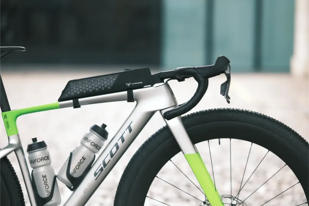 Una bicicleta Scott de color plateado y verde, con dos bidones y un dispositivo rectangular negro montado en el tubo superior, equipada con ruedas de 32”, está aparcada sobre una superficie de piedra.