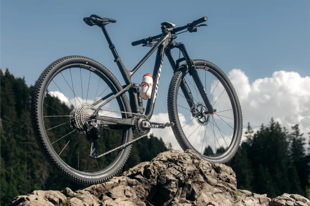 Una bicicleta de montaña con ruedas de 32 y un bidón acoplado se encuentra sobre una superficie rocosa al aire libre, con árboles y un cielo parcialmente nublado al fondo. Esta imagen refleja la revolución necesaria en el sector de los aficionados al ciclismo que buscan tanto rendimiento como innovación.