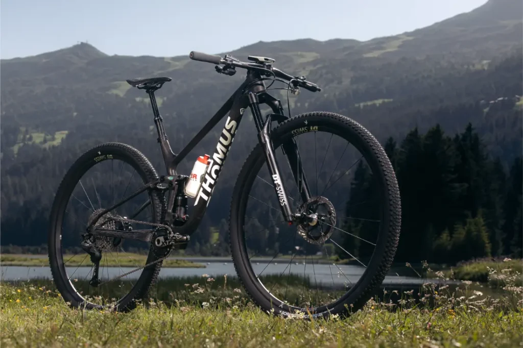 Una bicicleta de montaña Thömus negra con ruedas de 32 está aparcada sobre la hierba cerca de un lago, con el bosque y las montañas al fondo bajo un cielo despejado: una escena perfecta que refleja la revolución de las ruedas que está teniendo lugar en la industria ciclista.