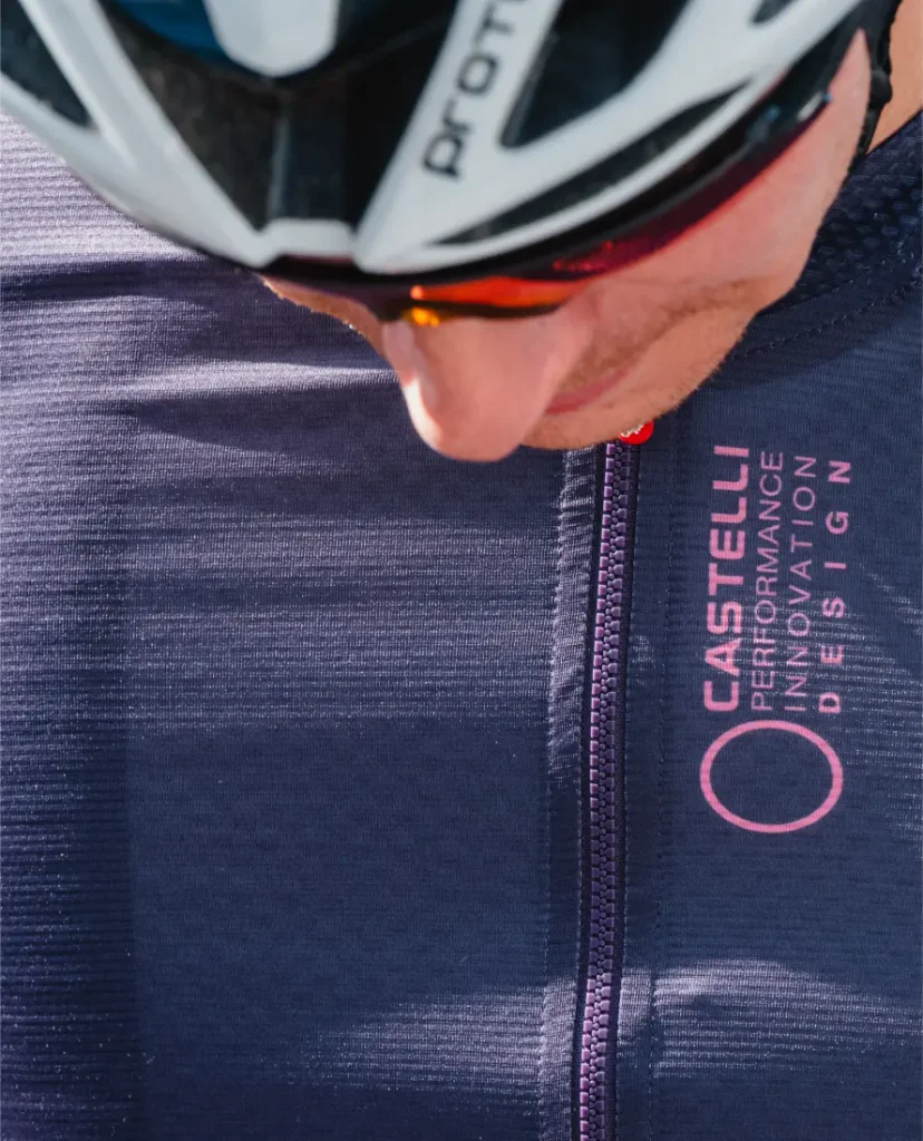 Primer plano de un ciclista que lleva casco y un maillot oscuro Castelli Corretto LS, un modelo de manga larga con el emblemático logotipo rosa y la inscripción «Performance Innovation Design».