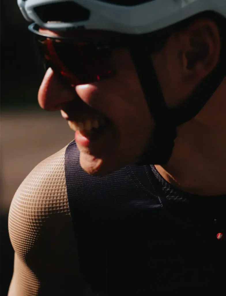 Primer plano de una persona que lleva un casco de bicicleta, gafas de sol y una camiseta sin mangas de malla «Castelli Transition», sonriendo y mirando ligeramente hacia un lado, bajo luz natural.
