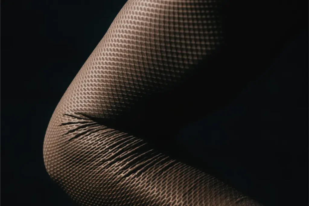 Primer plano de una rodilla doblada con medias de rejilla texturizadas, en las que se aprecian el estiramiento y los pliegues del tejido sobre un fondo oscuro, lo que recuerda a la elegancia ajustada propia de prendas de manga larga como el Corretto LS Jersey o el Castelli Transition.