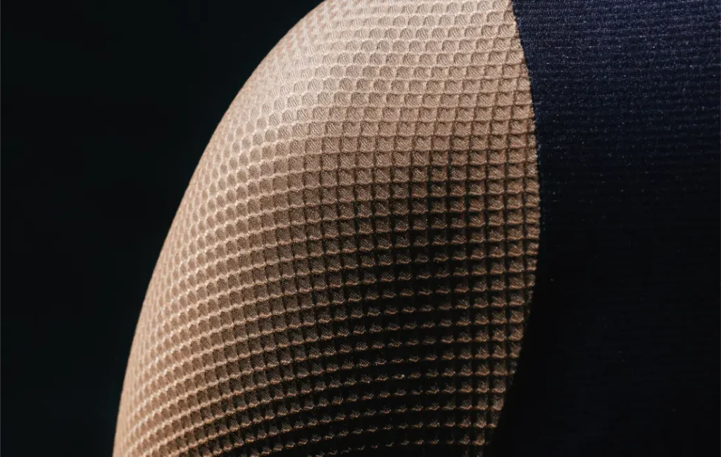 Primer plano del hombro de una persona que lleva una prenda de tejido beige con textura y diseño de panal de abeja de la colección Corretto LS Jersey, combinada con un trozo de tejido negro liso sobre un fondo negro.