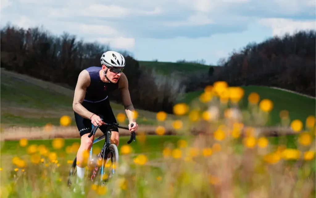 Un ciclista que lleva un casco Castelli Transition y unas gafas de sol recorre en bicicleta un paisaje cubierto de hierba con flores silvestres amarillas en primer plano, vestido con un maillot Corretto LS de manga larga.