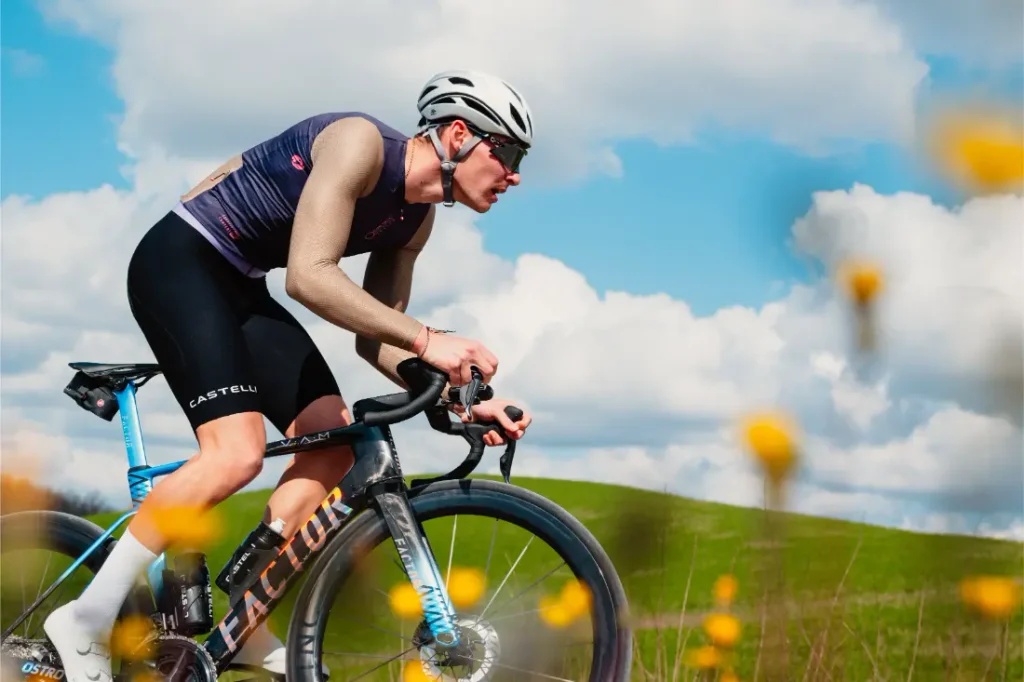 Un ciclista que lleva casco y gafas de sol monta en bicicleta de carretera al aire libre, vestido con un maillot Corretto LS de manga larga. En el fondo se ven colinas verdes y un cielo parcialmente nublado, mientras que en primer plano se aprecian flores amarillas.