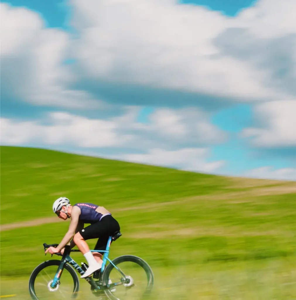 Un ciclista con equipación de competición, que lleva un maillot Corretto LS, pedalea a gran velocidad en una bicicleta de carretera por un paisaje cubierto de hierba, bajo un cielo azul con nubes dispersas.