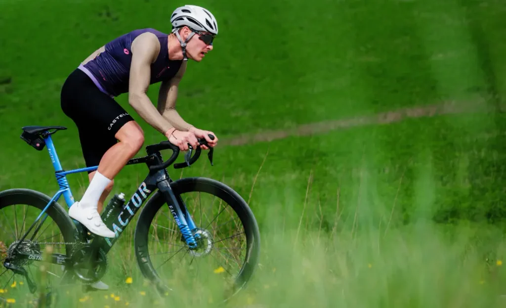 Un ciclista con equipación de competición —que luce un maillot Corretto LS y una camiseta de manga larga Castelli Transition—, pedalea cuesta arriba en una bicicleta de carretera azul por un paisaje cubierto de hierba, con un casco blanco y gafas de sol.