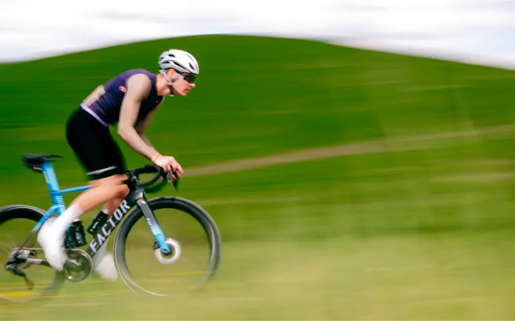 Un ciclista, con casco y gafas de sol, pasa a toda velocidad en su bicicleta de carretera junto a una colina cubierta de hierba, de un verde difuminado, bajo un cielo nublado, luciendo el elegante maillot Corretto LS de la colección «manga larga» de Castelli.