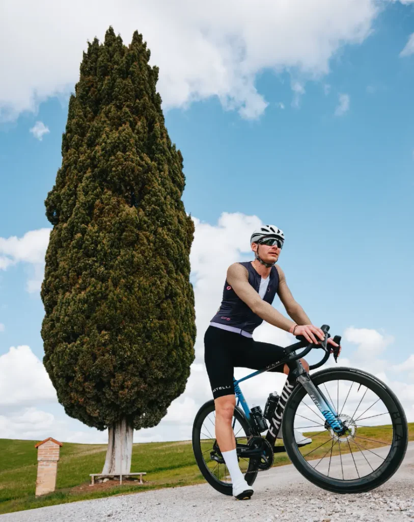 Un ciclista con gafas de sol y casco se detiene en su bicicleta de carretera frente a un ciprés alto, bajo un cielo azul con nubes dispersas, luciendo el maillot Corretto LS para disfrutar de una comodidad óptima en este recorrido panorámico.