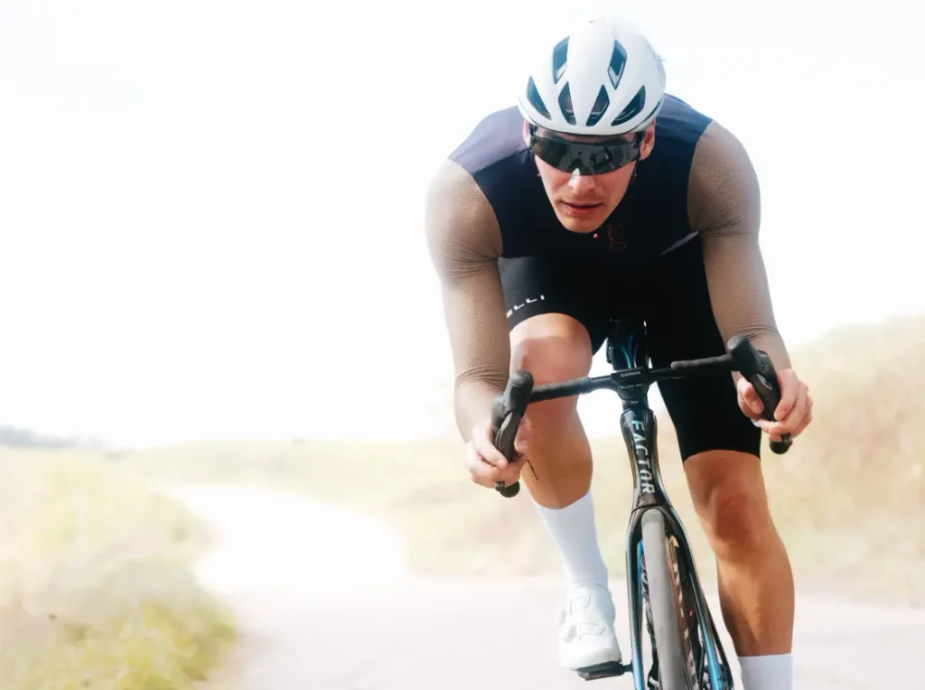 Un ciclista que lleva casco, gafas de sol y un maillot de manga larga «Transition» de Castelli circula en bicicleta de carretera por un carril al aire libre durante el día.