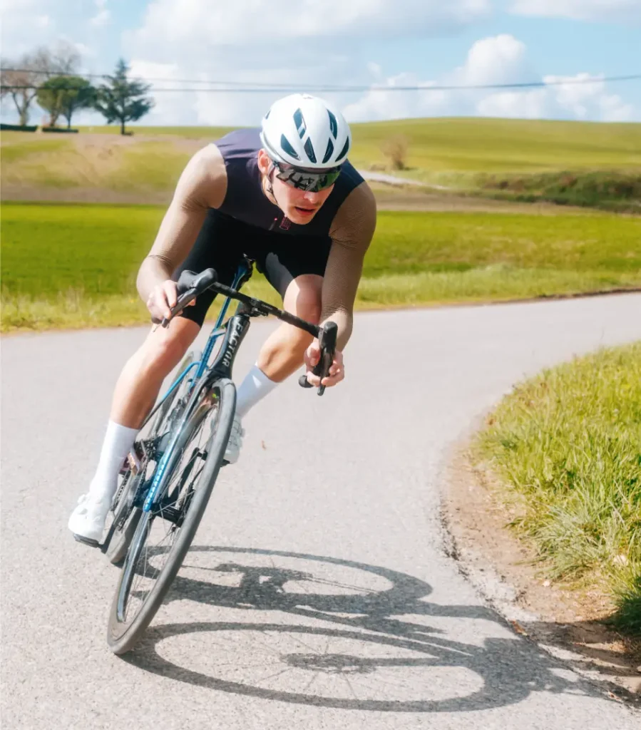 Un ciclista que lleva casco y gafas de sol, vestido con un maillot Corretto LS, se inclina al tomar una curva en una carretera rural rodeada de hierba y campos abiertos, bajo un cielo parcialmente nublado.