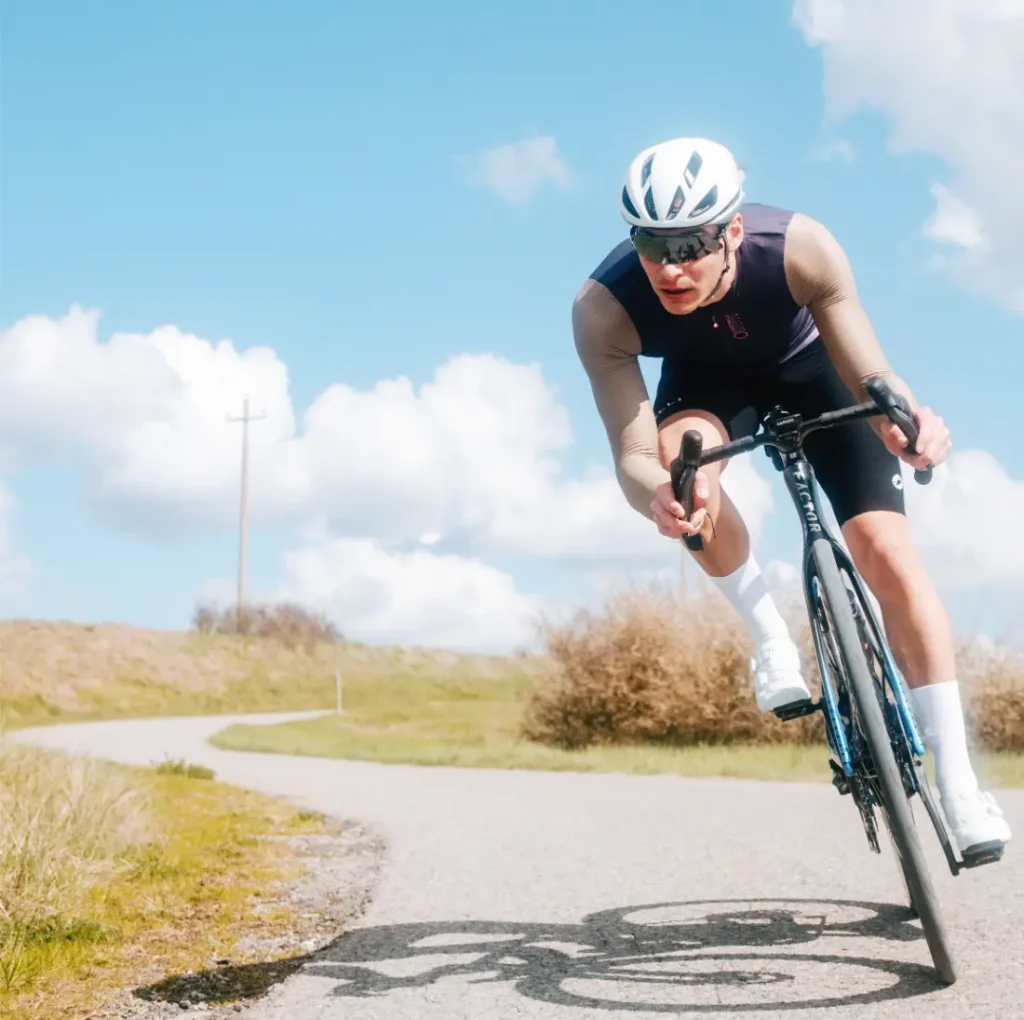 Un ciclista, con casco y gafas de sol, circula en bicicleta por una carretera sinuosa al aire libre, bajo un cielo azul con algunas nubes dispersas, luciendo el maillot de manga larga Corretto LS para disfrutar de una comodidad óptima.