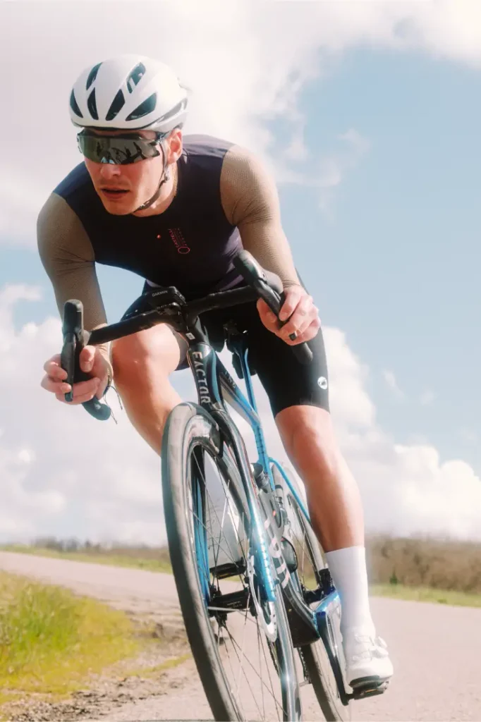 Un ciclista que lleva casco, gafas de sol y ropa de ciclismo, incluido el maillot Corretto LS, circula en bicicleta de carretera por un camino asfaltado bajo un cielo parcialmente nublado.