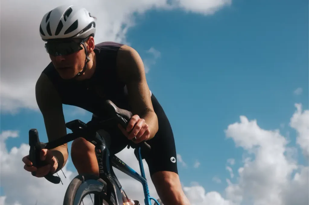 Un ciclista que lleva un casco blanco, gafas de sol y un maillot Corretto LS pedalea en una bicicleta de carretera al aire libre bajo un cielo azul parcialmente nublado.
