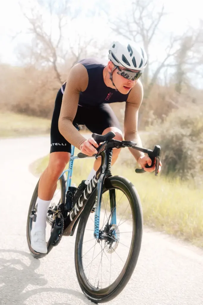 Un ciclista que lleva casco, gafas de sol y ropa de ciclismo —concretamente el maillot Corretto LS (manga larga) de Castelli Transition— circula en una bicicleta de carretera por un camino asfaltado con árboles y césped al fondo.