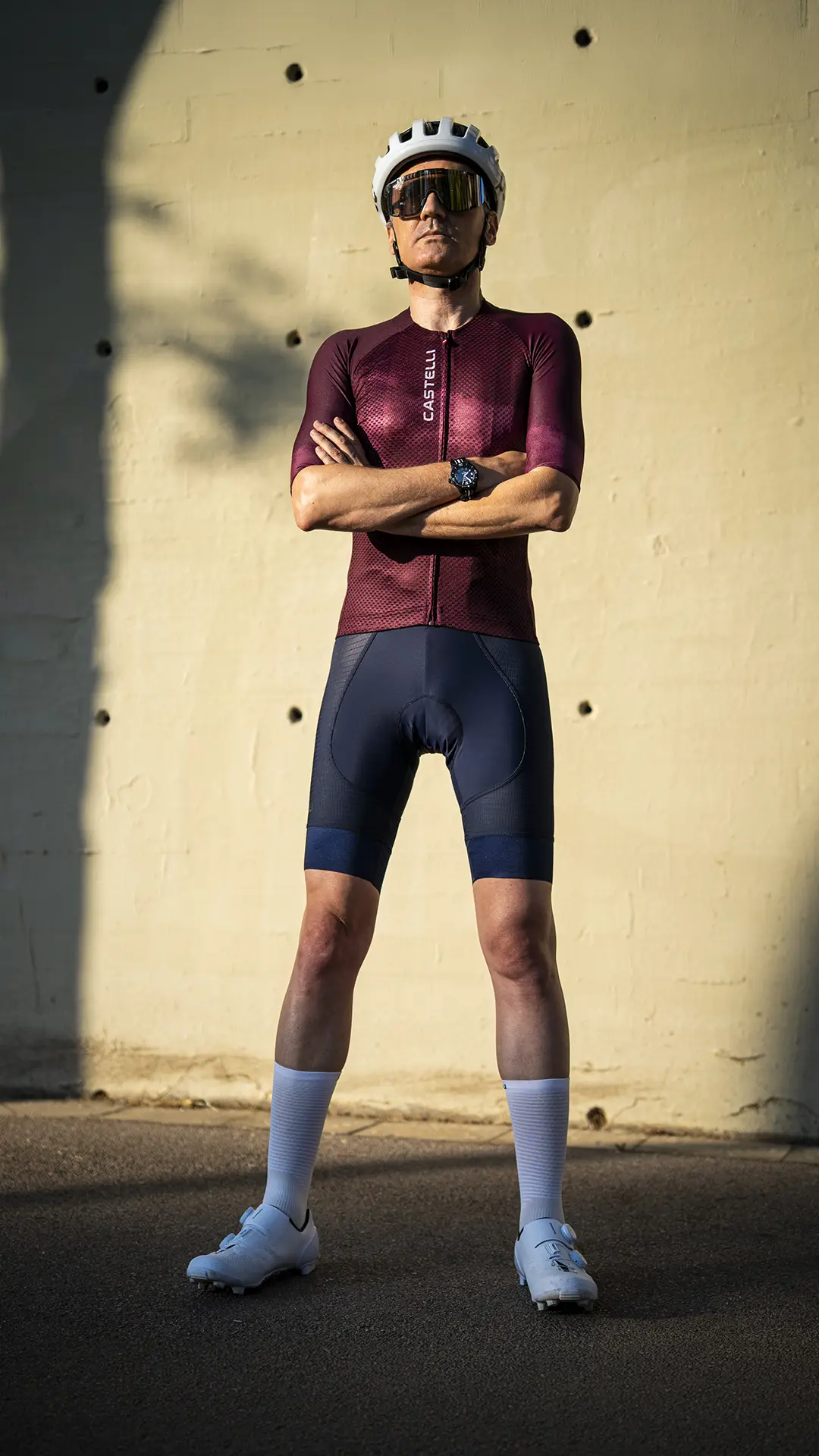 Una persona que lleva casco, gafas de sol, un maillot de ciclismo Castelli Climber’s A/C, unos pantalones cortos tipo culotte A/C, calcetines blancos y zapatillas de ciclismo posa con los brazos cruzados frente a una pared iluminada por el sol, mostrando su rendimiento en condiciones de calor con una confianza serena.