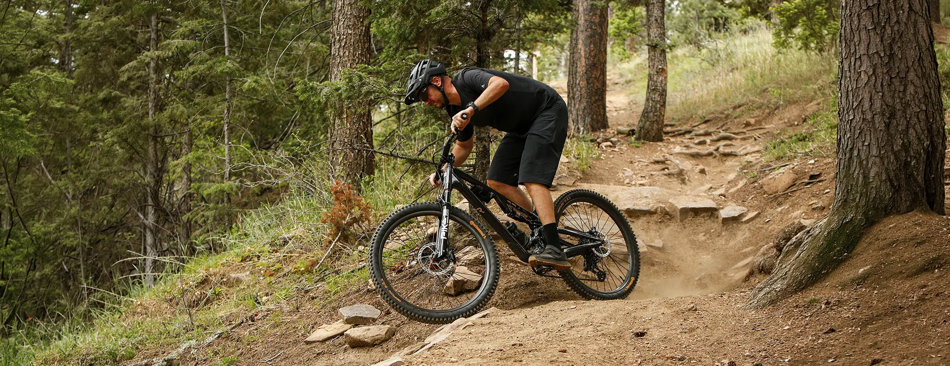 Una persona vestida con ropa de ciclismo negra va en bicicleta de montaña, equipada con cubiertas Continental Gravity para MTB, por un sendero de tierra en una zona boscosa, levantando polvo y esquivando rocas y raíces de árboles mientras explora nuevas combinaciones en un terreno exigente.
