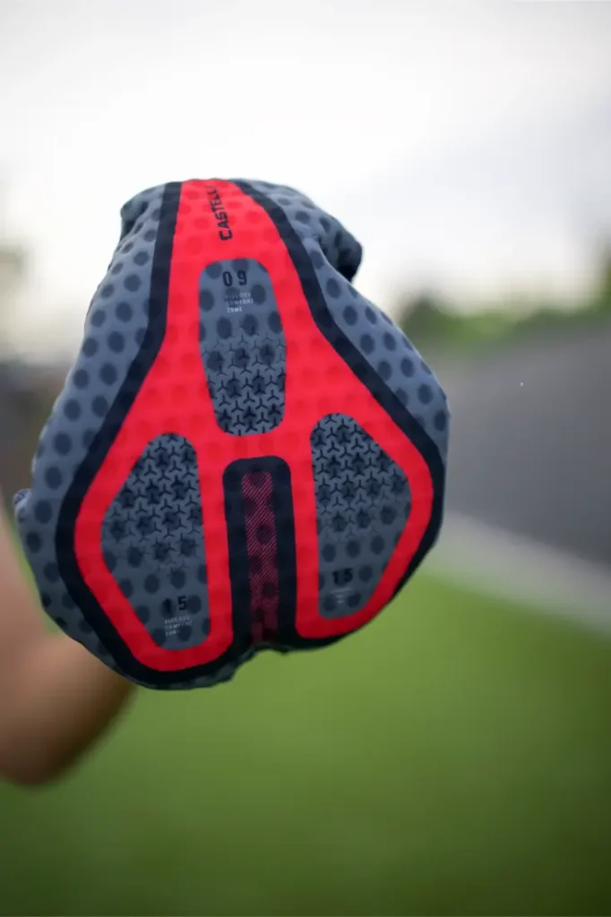 Una mano sostiene un guante de ciclismo acolchado, de color gris y rojo, con una empuñadura texturizada con motivos de estrellas, al aire libre, con la hierba difuminada al fondo; perfecto para combinarlo con el conjunto Castelli Climber’s A/C y mejorar el rendimiento térmico en las salidas en días cálidos.
