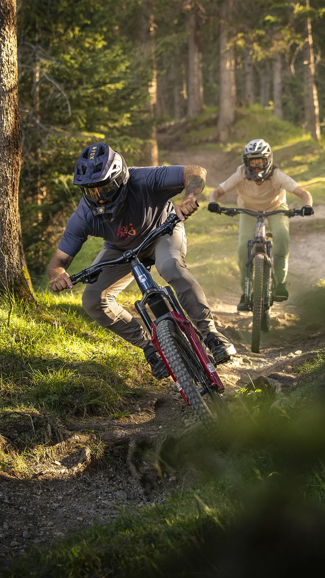 Dos personas con casco bajan en bicicletas de montaña eléctricas JAM e-MTB 2027 por un sendero de tierra en un bosque, mientras la luz del sol se filtra a través de los árboles.