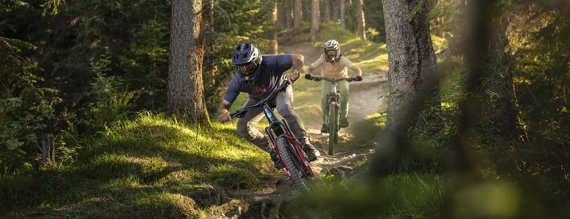 Dos personas con casco bajan en unas bicicletas de montaña eléctricas Gama FOCUS e-MTB 2027 por un sendero de tierra que atraviesa un bosque, mientras la luz del sol se cuela entre los árboles.