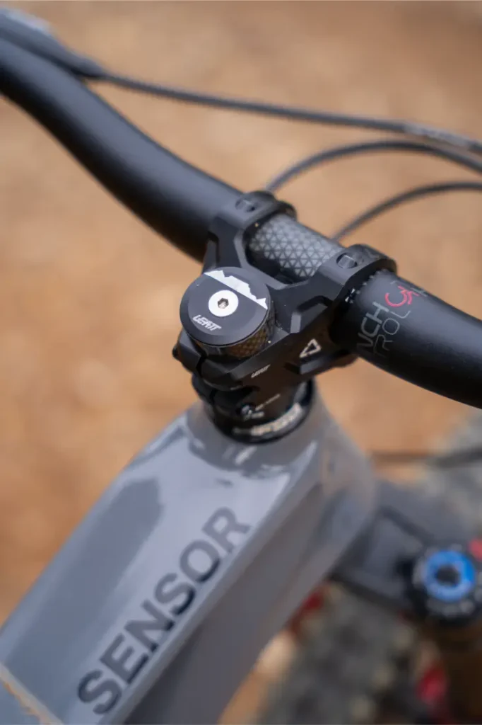Primer plano del manillar y la potencia de una bicicleta, equipados con puños ReaFlex 6.0 que ofrecen un agarre y un control superiores. La marca «SENSOR» es visible en el cuadro de la bicicleta, mientras que el manillar Leatt Launch Control 6.0 combina a la perfección con una tapa superior «NSB» en la potencia Gravity 6.0, lo que le confiere un aspecto elegante y de alto rendimiento.